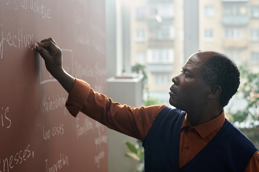 Side view portrait of Black college professor writing on chalkboard while preparing for lecture in college