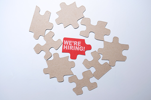 Piece of a jigsaw puzzle with We're Hiring text over a white background. Human resources management concept