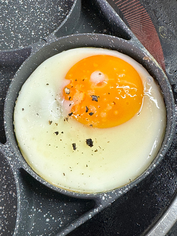 Stock photo showing fried egg that is being cooked in a stainless steel, non-stick frying pan on a ceramic hob. The pan has rings to crack eggs into in order to keep them separate and stop the egg whites from merging.