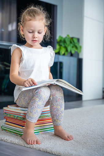 Little girl is sitting on stack of children's books and leafing through a book with fairy tales in home living room. High quality photo