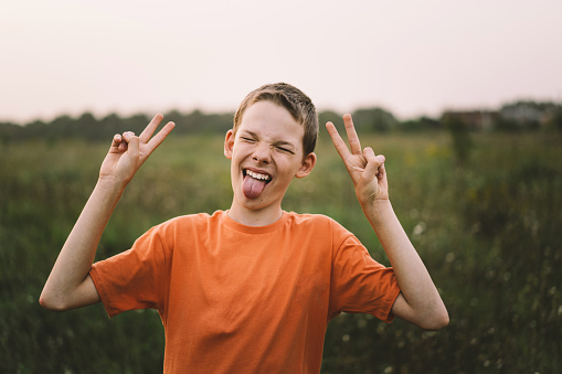 Portrait of a funny boy shows two fingers, victory or peace. Funny boy in a orange T-shirt playing outdoors on the field at sunset. Happy child, lifestyle.