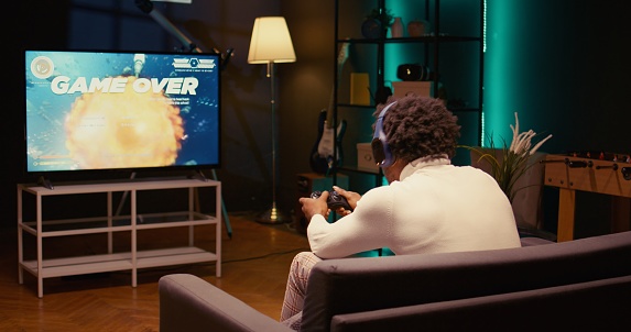 Gamer receiving game over screen on ultrawide smart TV, losing all health, failing to win level and beat enemies. BIPOC player defeated in science fiction singleplayer videogame, panning shot