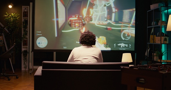 Man watching gameplay footage of videogame competition on gigantic smart TV, relaxing on couch. BIPOC gamer in home theatre following gaming tournament on television set, zoom out shot
