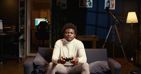 Man focused on playing singleplayer videogames on TV in cozy living room. BIPOC gamer using controller to beat science fiction shooter game on gaming console, relaxing on couch