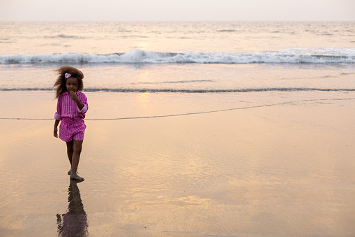 Charming African-American girl with flowing hair walks along sand by water, enjoying cool waves washing her feet .Happy childhood. Positive children's emotions. Summer holidays. Recreation .
