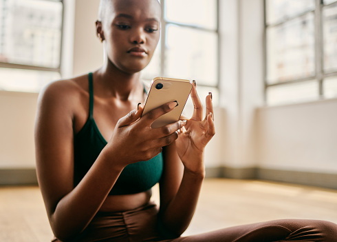 This stock image captures the serene ambiance of a woman fully immersed in her post-yoga routine. After a rejuvenating yoga class, she finds a moment of tranquility as she sits cross-legged, gently texting on her mobile phone. The image portrays the perfect balance between mindfulness and modern connectivity, showcasing how technology can seamlessly integrate into our lives even after moments of self-care and inner peace.