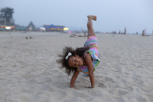 6-7 years old African-American girl does gymnastic somersault on seashore. Girl is gymnast. Child plays sports on beach. Harmonious development of children. Children's holidays. Happy childhood.