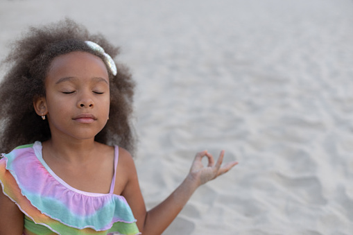 Children's yoga meditation. Portrait of African-American girl with curly hair who is sitting on beach summer day. Children's holidays. Harmonious development of children. Sports and health.