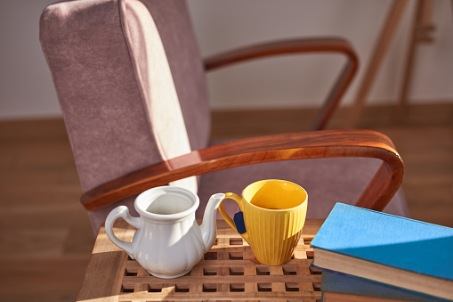 Teapot and tea cup with books and a comfortable chair in a sunny home interior.