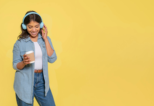 a happy dancing woman with blue  headphones  in jeans and denim shirt drinking coffe on yellow background with copy space do what you like