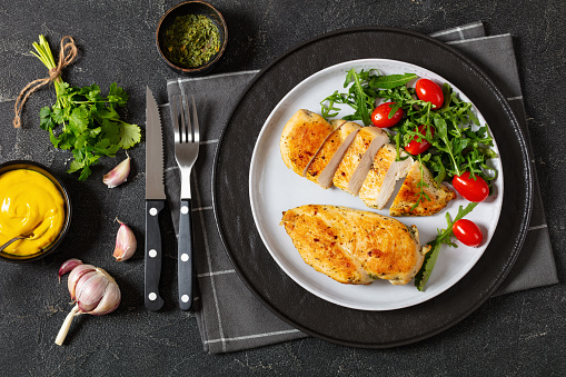 overhead view of juicy fried chicken breasts with mixed green salad, tomatoes on plate on grey concrete table with cutlery and yellow mustard, flat lay