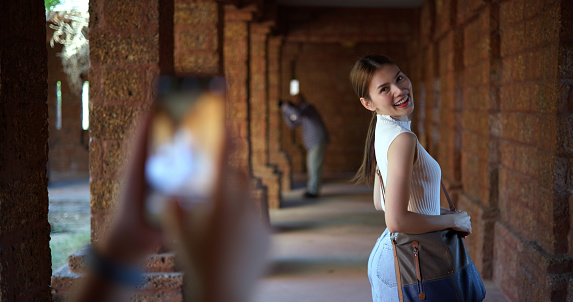 Selective focus, Beautiful woman pose smiling with happiness at archaeological site, Young female local guide use smartphone to takes photos young tourist while visit ancient architecture
