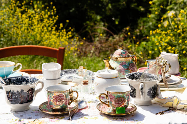 afternoon tea, dessert table, cake, smashed cake, fruit plate, grapes, raspberries, macarons, cookies, teapots, mugs, outdoor party, vintage crockery, floral tablecloth, cheese board, crackers, sliced cheese, food arrangement, meringue, roses, layered desserts, celebration, food styling, spring garden, table setting, books as riser, colorful chair, vibrant display, sweet treats, jam, sweets, lemon slice, strawberry, artistic tea setup, banquet, outdoor feast, decorative table, gathering, social dining, celebration spread, eclectic mix, rustic elegance, floral decoration, food and drink setup