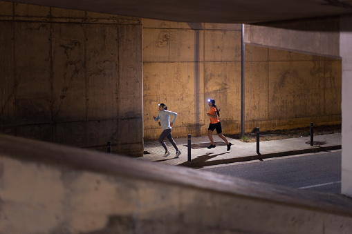 Two young female athletes, displaying focus and determination, run with intensity outdoors in the city at night, illuminated by headlamps as they undergo rigorous training. Wide shot with copy space