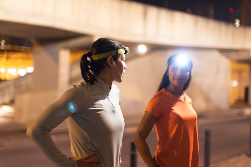 In the city night, two elated young female athletes converse happily, sharing smiles and camaraderie after a demanding training session while utilizing headlamps.