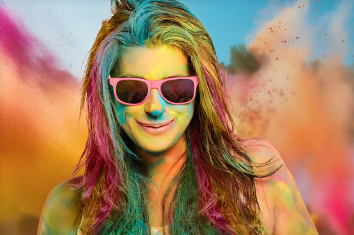 Carefree cheerful woman covered in rainbow colored powder celebrating holi color festival. Young woman having fun with colorful powder outdoors. Closeup portrait with copy space
