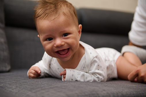 Authentic portrait of a cute Caucasian baby boy lying on tummy, playing, crawling on couch at home, smiling cutely and happily looking at camera, wearing white body and diaper. Free copy ad space