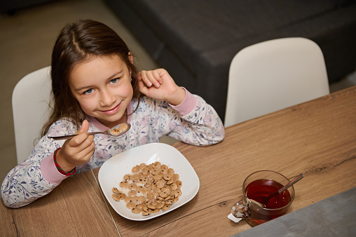View from above Caucasian adorable little child girl cutely smiling looking at camera, sitting at table and taking her delicious healthy breakfast. Cute kid eating flakes cereals in the home kitchen