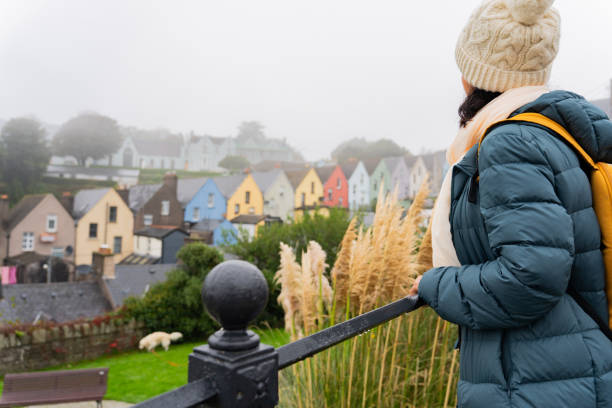 istock Unknown woman with a knit hat and scarf leaning out from a railing while enjoying a beautiful view of the colorful houses of Cork on a foggy day on her trip through Ireland by car 1980509351