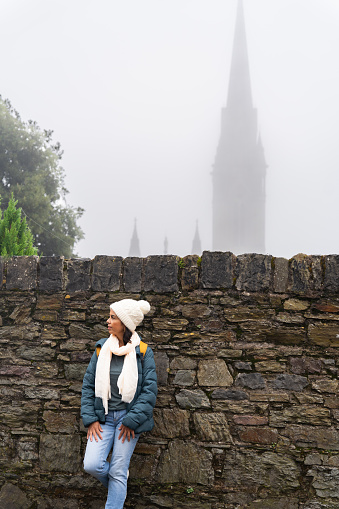 Latin woman enjoying her cultural visit to a small town near Cork where we can see a cathedral through the fog while she enjoys the views and the silence of village.