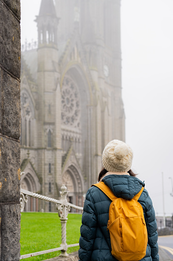 Unknown woman with winter hat and backpack enjoying splendid view of the cathedral on a cloudy day near Cork in Ireland