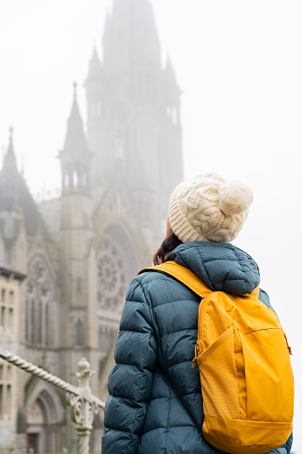 Close up of female traveler dressed in winter clothes and wool hat and backpack while looking in amazement at the cathedral of a town near Cork in Ireland on a foggy day