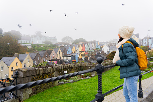 Unrecognizable woman enjoying beautiful views from the top of colorful houses and birds flying in a picturesque and striking town near Cork on her road trip through Ireland on a foggy day
