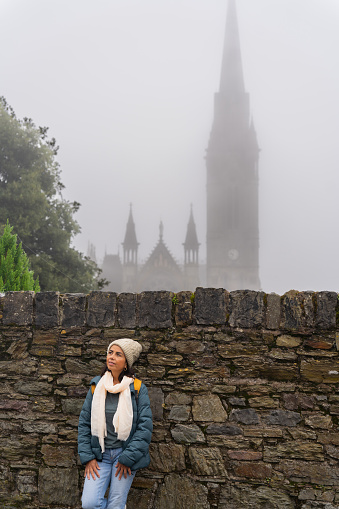 Latin woman leaning on a large natural stone wall with a large cathedral in the background being able to see in the thick fog in a town near Cork in Ireland