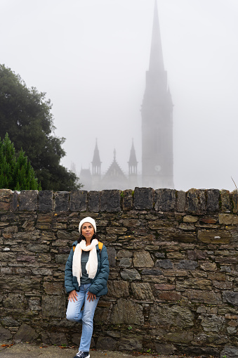 Latin woman leaning on a stone wall being able to see behind a large cathedral hiding in the thick fog in a town near Cork in Ireland