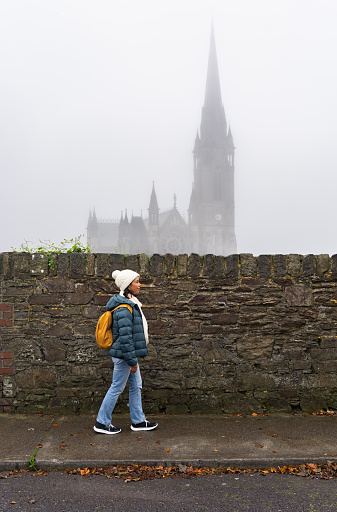 Traveler woman with a backpack and winter hat walking next to a large stone wall with a cathedral seen in the fog in the background