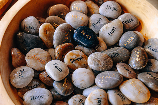 A pile of colorful engraved stones on a wooden bowl with a different word painted on each. Peace, Hope, Dream, Trust, and Believe on River Rocks for inspiration
