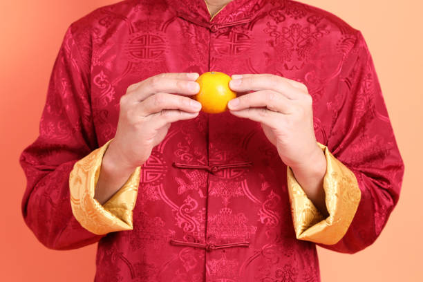 Happy Chinese new year. Hand of asian chinese senior man wearing red traditional clothing with gesture of hand holding orange isolated on orange background.
