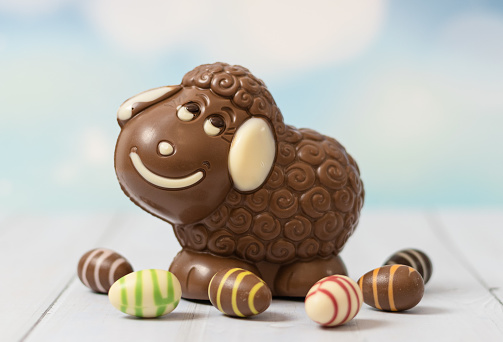 One cute chocolate happy lamb with easter candy eggs around it stands on a white wooden table, close-up side view.