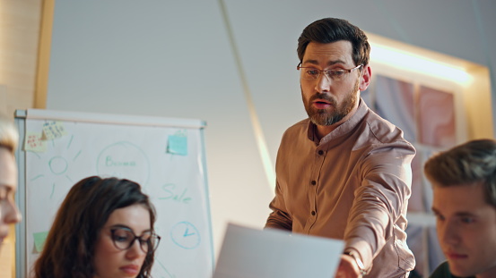 Focused bearded manager supervising work business team at corporate meeting close up. Serious man boss explaining project strategy to diverse group coworkers in boardroom. Ceo looking design samples.