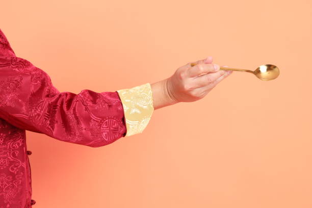 Happy Chinese new year. Hand of asian chinese senior man wearing red traditional clothing with gesture of hand holding golden spoon isolated on orange background.