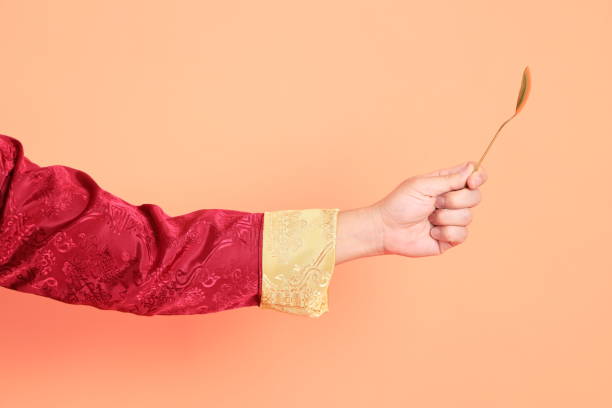 Happy Chinese new year. Hand of asian chinese senior man wearing red traditional clothing with gesture of hand holding golden spoon isolated on orange background.