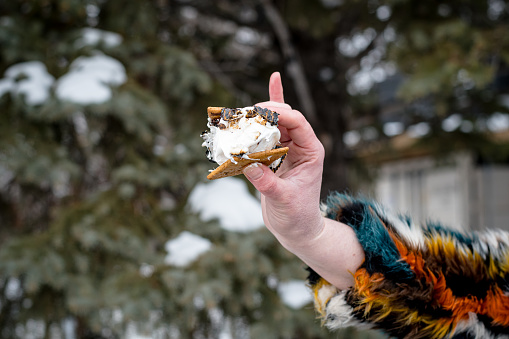 Indulging in winter sweetness, the nonbinary individual takes a delightful bite of their crafted s'mores, a moment of pure bliss by the fireside.