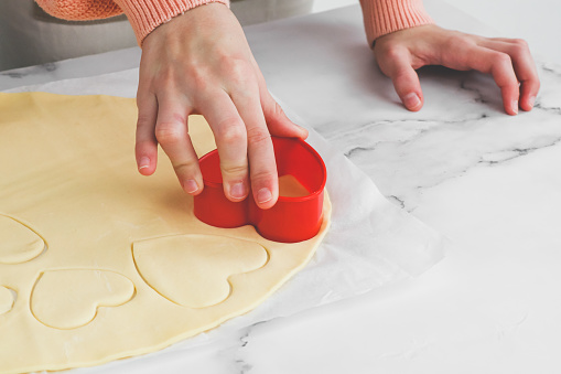 The hands of a caucasian teenage girl cuts out a shortbread dough with a red plastic heart shape on a white parchment on a marble table, close-up side view.The concept of homemade baking and preparation for valentine's day.