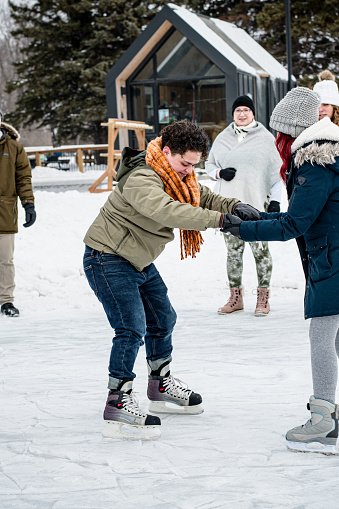 With cautious courage, the trans man takes his first steps on the ice, surrounded by the warmth of chosen family, creating a scene of shared encouragement and joy in the winter air.