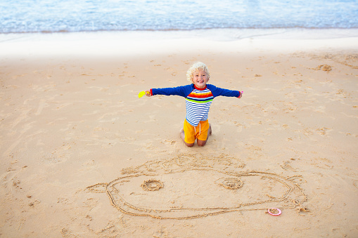 Child playing on tropical beach. Little boy drawing his portrait in sand at sea shore. Family summer vacation. Kid building sandcastle. Water and sand fun for children. Kids play on ocean coast
