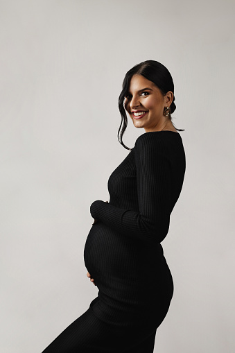 Portrait of a beautiful young pregnant woman wearing an elegant black dress holding a belly, studio shot in front of a white background