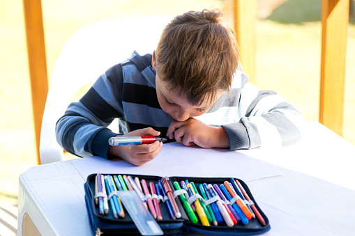 Boy about to draw on a blank paper with black marker. He is outdoors sitting at a table.