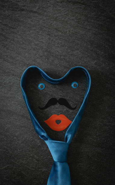One blue male tie with decorative eyes and a paper mouth smile lie in the center on a black stone background with copy space on the sides, flat lay close-up. Valentine's day concept.