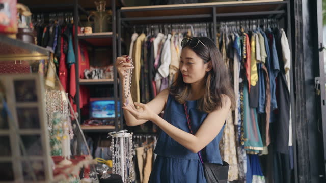 Asian woman looking through clothing and picks up a necklace  holding it against herself to try.