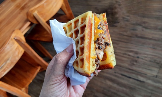 Hand holding waffle with toppings of beef slices, mushrooms, melted cheese, mozzarella and onion.