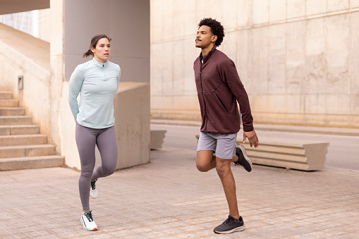 Focused and resolute, a young woman and man, dressed in sportswear, perform meticulous warm-up exercises together before embarking on their running training in the city. Wide shot with copy space