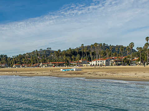 Santa Barbara, CA, USA - January 27, 2024: Stearns Wharf viewpoint. First blocks of West Cabrillo Blvd behind sandy west Beach under blue cloudscape. White facades, green palm trees, hills,,