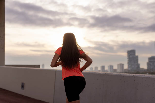 Running along a concrete bridge with city views during sunset, a young adult female runner in sportswear, captured from behind, exerts significant effort in a sprint. The scene depicts a moment of intense and hard training, highlighting the runner's determination and commitment to pushing her limits.