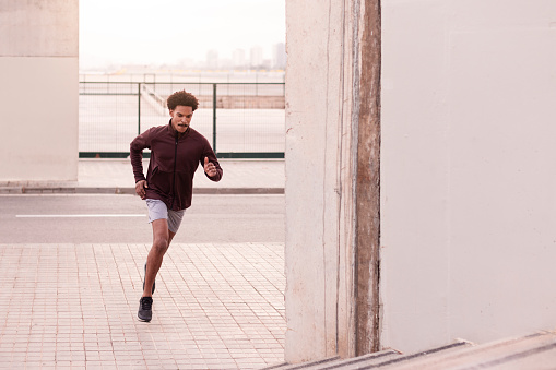 During sunset, outdoors in the city a young male athlete sprints with intensity during a running training, heading towards a concrete staircase. His determined and concentrated expression underscores a commitment to achieving better results, highlighting the intensity of the workout. Wide shot with copy space.