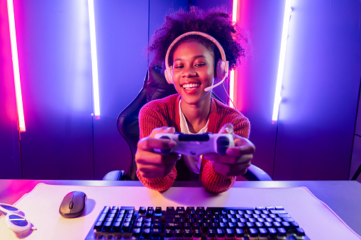 Host channel of gaming streamer, African girl playing online game with joystick, talking with viewers media online on microphone. Esport skilled team players in neon color lighting room. Tastemaker.